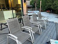 Four patio armchairs shown on a wooden deck near a house, angled view showing seat and back design.