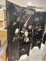 Four-panel black lacquer screen with stone and jade carved inlay, birds and floral motifs with gold trim on one side, shown in an indoor storage setting.