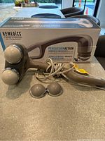 HoMedics percussion action handheld massager displayed outside its box with two extra interchangeable massage heads and power cord visible.