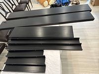 Photo displaying multiple Ikea black and brown wall shelves laid flat on floor, including larger and smaller shelves showing length and condition.