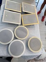 Full view of 4 rectangular white JBL wall/ceiling speakers and 4 round white Polkaudio RC60i speakers laid out on a table showing frame color and shape.