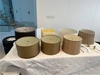 All 7 lamp shades and 2 finials arranged on white quilted surface, showing round shades in front and back, square yellow shade in middle, two metal finials to right.