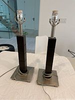 Two modern table lamps side by side on a white cloth. Each lamp has a square stainless steel base, espresso colored stacked wood block stem, and metallic lamp socket without shade.