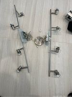 Photo showing both stainless steel track lighting sets side by side on a floor, displaying the four adjustable spotlights on each track and circular mounting bases with exposed wiring.