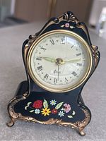 Front view showing clock face with Roman numerals, ornate gold trim, and floral painted decoration on black body.