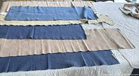 Six washable table runners laid out on a surface; three are blue and three are taupe with scalloped edges, showing varying fabric textures and patterns.
