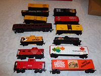 Photo showing 13 HO scale model railroad cars arranged on a cloth surface. Visible variety of colors and branding including CP Rail yellow boxcar, Canadian Pacific red cabooses and passenger car, Gaine's Gravy Train boxcar, Canadiana 1867 boxcar, and a black tanker car.