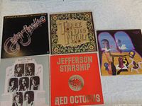 Four vinyl LP records arranged in two rows: top row has Captain & Tennille Greatest Hits (black cover, stylized script), Three Dog Night with ornate border cover, and a third album with stylized number 3 and characters. Bottom row shows Jefferson Starship Red Octopus (red cover with octopus) and a Three Dog Night album with black and white portraits on cover.