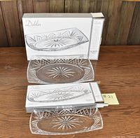 Two Dublin Crystal serving trays placed on a wooden surface with their original boxes displayed behind each tray. Each tray is clear crystal with cut starburst patterns, oval shaped.