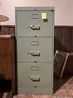 Green 3-drawer metal filing cabinet against a light brown wall with a wooden piece of furniture adjacent on the left and some framed art on the floor to the right. Cabinet is rectangular and each drawer has a handle and lock.