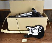 Photo showing a black and white wired guitar controller, silver wired microphone, and black drum pads inside a cardboard box on wooden floor.