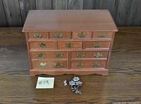 Full view of wooden jewelry box with drawers and two brooches in front on a wooden surface.
