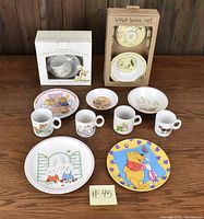 Image shows an assortment of children's tableware including unopened boxed feeding sets, plates, bowls, and mugs with various character designs such as Guess How Much I Love You, Peter Rabbit, and Winnie the Pooh.