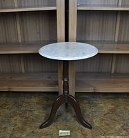 Photo of a small round table with white marble top and dark wooden pedestal base with three curved legs, placed in front of wooden shelves.
