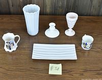 All items displayed on wooden surface: milk glass pitcher, vase, candle holder, wine glass, white serving tray, and two commemorative mugs.