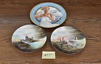 Top view of three decorative plates, one depicting Helen of Troy with blue and gold border, two others with coastal scenes.