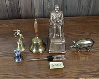 Photo showing entire lot on a wooden surface including three brass bells, a seated statue, two pig figurines, and a letter opener case.