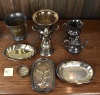 Photo of all miscellaneous silver plated dishware arranged on a wooden surface. Items include three trays (two oval, one rectangular with decorative raised design), a dinner bell, small candy dish, teapot, two ice/champagne buckets, and a wine pitcher.