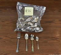 Plastic bag filled with a variety of metal spoons. On the wooden surface in front rest four pieces of silverware: one fork and three spoons of increasingly smaller sizes.
