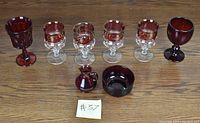 Photo showing 4 Kings Crown Thumbprint Ruby red wine glasses with clear stems, 2 clear stem glasses with ruby bowls, 1 ruby goblet, 1 small ruby pitcher, and 1 ruby bowl arranged on wooden surface.