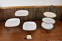 Two sets of serving plates with metal holders shown on wooden table against wood-paneled wall. One set has white square plates on separate scrollwork stands; other set has three oval cream and gold plates on single vertical holder.