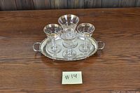 Four wine glasses with gold rims (three small, one large) placed on a silver serving tray on a wooden surface.