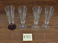 Four tall sling glasses arranged in a row on a wooden surface. Each glass features an etched cornflower pattern. One glass has a red stem and base, while the other three have clear stems and bases. A sticky note with '#42' is visible in front of the glasses.