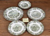 Five Staffordshire plates laid out on table, showing hand engraved scenic illustrations with floral borders in various sizes.