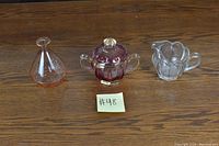 Photo shows three glassware items on wood surface: a clear glass vase, a purple-tinted sugar bowl with lid and handles, and a clear glass creamer with handle.