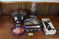 Photo of four kitchen appliances including Rival Crockpot slow cooker, Ninja personal blender, Cuisinart Griddler contact grill, and boxed Pro Smart Slicer, all placed on wooden table.