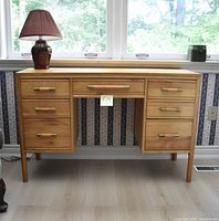 Front view of light brown wooden desk with brass-tone handles, seven drawers, tapered legs
