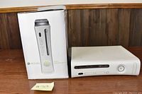 White Xbox 360 console shown next to original retail box on a wooden surface, highlighting front of console with power button, disc tray and two USB ports.