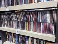 Two rows of assorted music CDs in jewel cases on shelf, titles of many artists and albums visible but not fully legible in the image