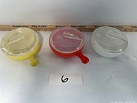 Three Glassbake bowls in yellow, red, and white, each with a clear glass lid on top, arranged in a row with measuring ruler behind them for scale.