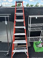 Full view of the orange fiberglass step ladder standing upright outdoors with a black backdrop and other items visible beside it.