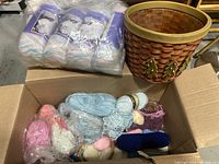 Photo showing a cardboard box filled with assorted skeins of yarn and a plastic-wrapped pack of pastel multicolor yarn. The woven waste basket with Christmas tree decorations is beside the box.