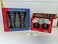 Two boxed Philadelphia Flyers collectible cups and boxed Budweiser pint glass set with Budweiser American Ale beer tap handle behind, on a white surface with measuring tape in background.