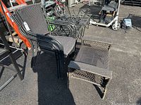 Four black stacking outdoor chairs with woven synthetic seating and metal frames alongside one wicker ottoman, all placed on asphalt outdoors.