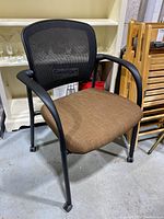 Front angle showing brown fabric seat, black mesh backrest, armrests, and rolling casters on floor.