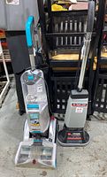 Wide view of the two Hoover floor cleaners standing side by side on a concrete floor with chairs in the background