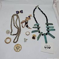 Full view of jewelry lot showing twisted rope chain necklace, sword chatelaine pin, green stone and black bead necklace, various pins and brooches including purple bow and circular rhinestone brooch.