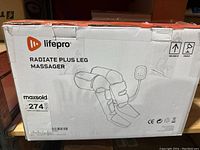 Photo of the sealed white and orange LifePro Radiate Plus Leg Massager box showing product name and schematic line drawing of the leg massager with wired remote control.