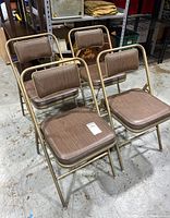 Four Samsonite metal folding chairs with brown wood grain vinyl padded seats and backs arranged in grouping on concrete floor.