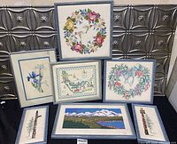 Photo showing seven framed needlepoint artworks including floral wreaths, a map, and a landscape.