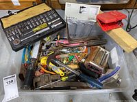 Bin filled with assorted hand tools including socket wrench set, hammers, pliers, screwdrivers, saw blade, red metal tool, and miscellaneous hardware, placed on wooden surface.