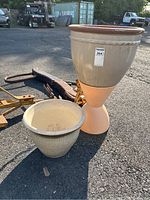 Two planters set on pavement; one tall planter with a peach-colored plastic base and beige ceramic upper part, and one smaller beige ceramic planter.