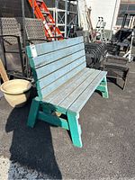 Side perspective of vintage blue wooden bench showing faded blue paint on slats and green wooden legs, placed on asphalt surface.