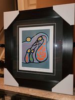 Photo of framed artwork "Family Teaching 1" by Bruce Morrisseau, showing the full frame with corner protectors and the print with colorful Indigenous-style figures.