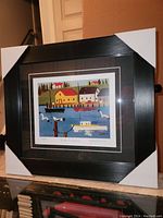 Framed Maud Lewis print leaning against a wall, showing the full frame and print with protective corner covers.