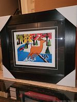 Photo showing the full framed artwork with protective corner covers on the black wood frame, displaying the stylized deer and colorful fall landscape.